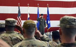 A military officer in flight suit salutes while standing at attention in front of four flags including the American flag and Air Force flags. The officer is positioned against a large American flag backdrop with red and white stripes. In the foreground, seated military personnel in camouflage uniforms and caps are saluting back.