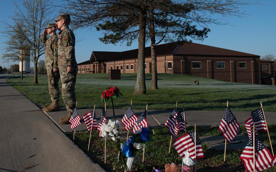 Two military members in camouflage uniforms salute while standing next to a patch of grass marked with flowers and small U.S. flags.