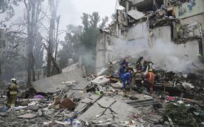 Rescue workers in blue and orange suits stand on top of a large pile of rubble and debris next to the facade of a damaged building.