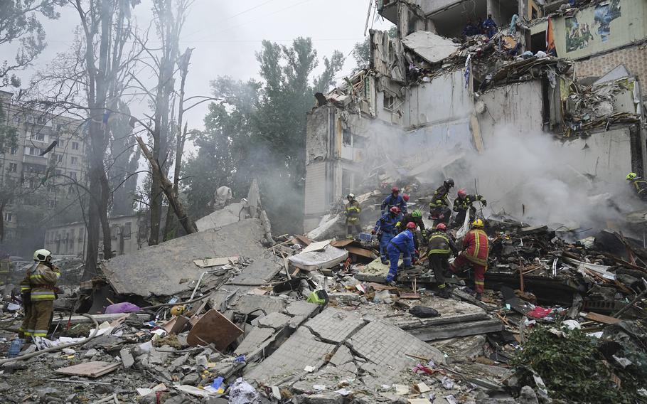 Rescue workers in blue and orange suits stand on top of a large pile of rubble and debris next to the facade of a damaged building.