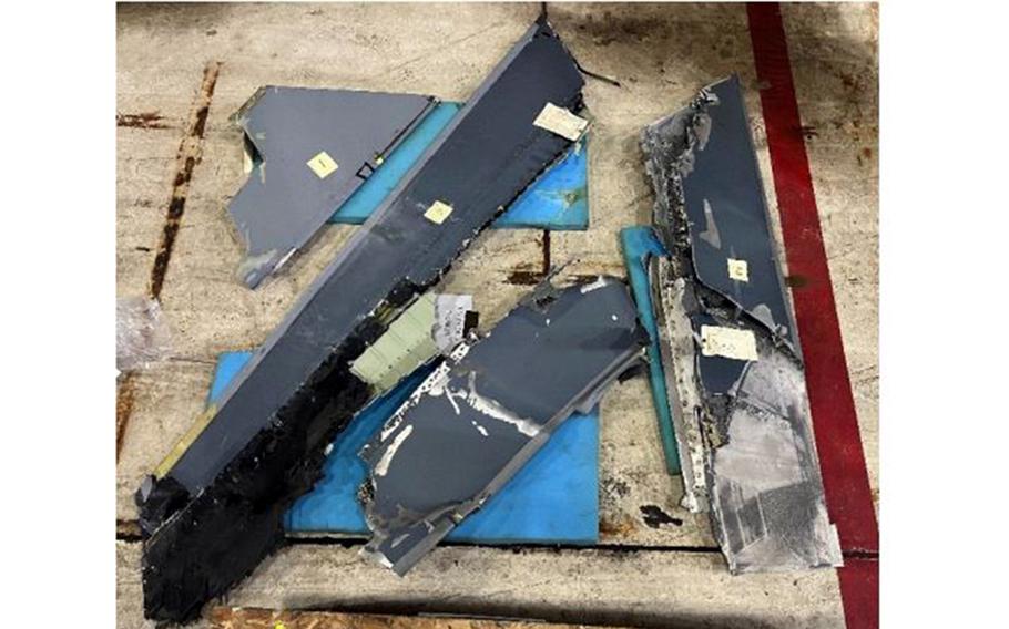 Wreckage of a military aircraft scattered on concrete ground, showing twisted gray and blue metal fragments, damaged panels and debris.