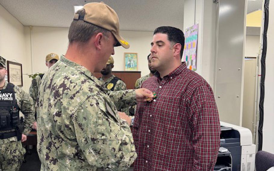 A Navy officer in camouflage uniform and hat pins a medal on the chest of another officer in a flannel civilian shirt.