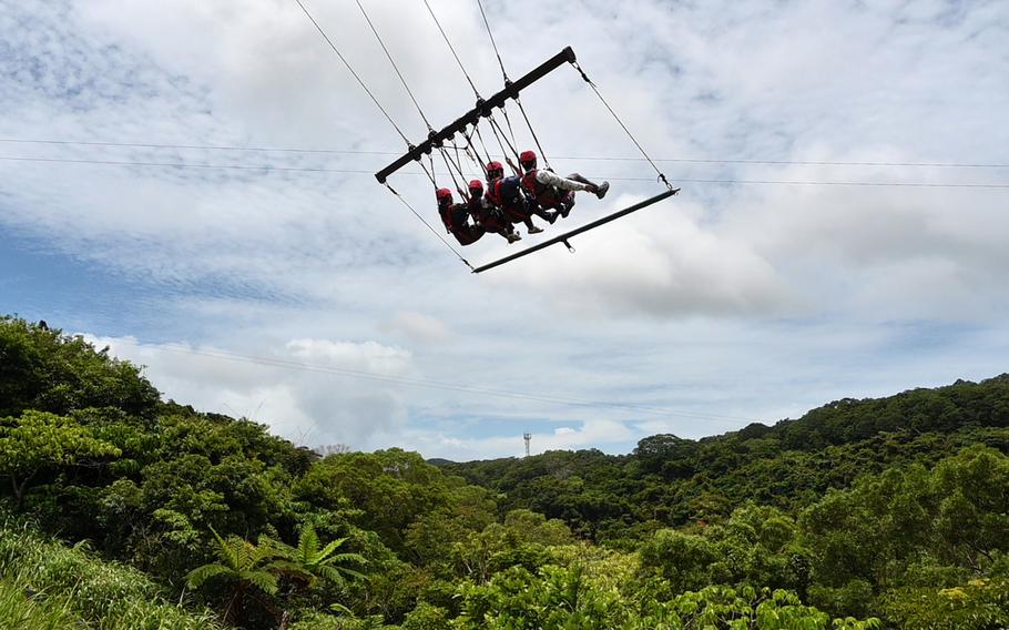 A group of people wearing red helmets are on a swing high above a dense green forest canopy, with rolling hills and a communications tower visible in the distance.