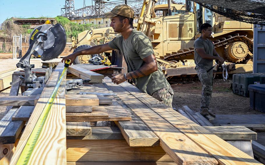A soldier in camouflage pants and a green t-shirt uses a measuring tape on a construction site. 