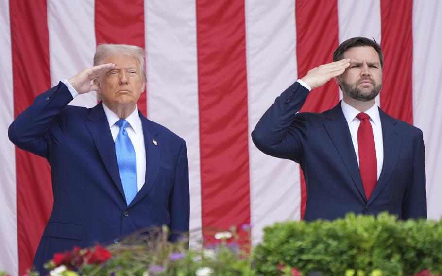Donald Trump and JD Vance, both wearing dark suits, stand and salute in front of a large U.S. flag.