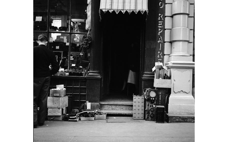A black-and-white, 1950s-era image of a man, seen from behind, standing on a sidewalk and looking through the front window of a shop.