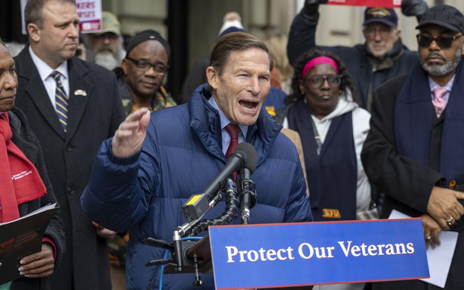 Blumenthal, wearing a blue coat, stands at a podium with a placard reading “Protect Our Veterans” and speaks into a microphone, with people standing behind him, some holding signs.