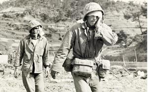 A black-and-white, 1940s-era image of two injured military members, one with his hand on his face, walking across a rocky beach to medical treatment.