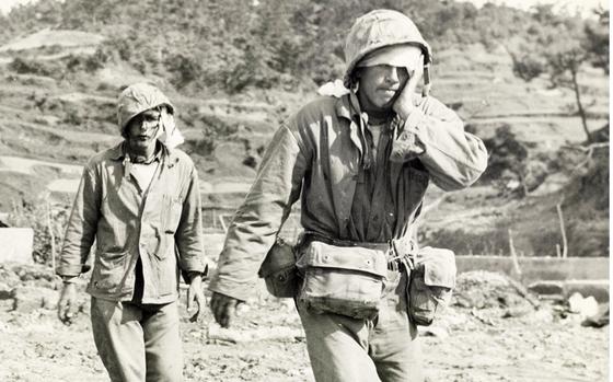 A black-and-white, 1940s-era image of two injured military members, one with his hand on his face, walking across a rocky beach to medical treatment.