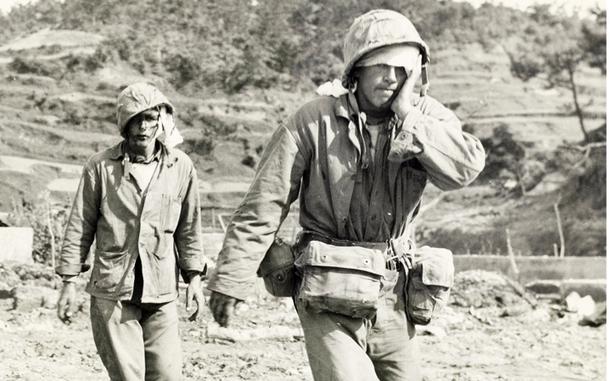 A black-and-white, 1940s-era image of two injured military members, one with his hand on his face, walking across a rocky beach to medical treatment.