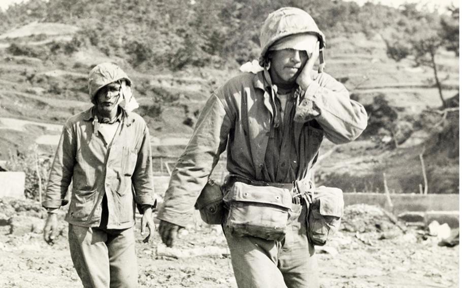 A black-and-white, 1940s-era image of two injured military members, one with his hand on his face, walking across a rocky beach to medical treatment.