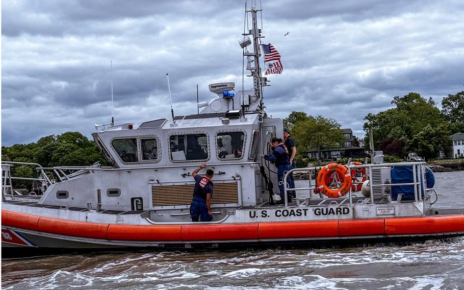 Agents in blue uniforms and life vests stand on the deck of a Coast Guard ship in open water with the tree line in the background.