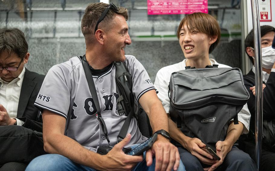An American man with a camera around his neck and a Japanese man with a bag in his lap sit together on a train, facing each other and smiling.