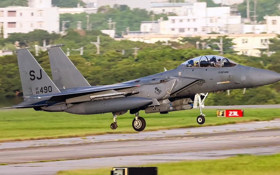 A gray F-15E Strike Eagle fighter jet comes in for the landing on a runway, with green grass in the background. While the landing gear is deployed, the right back wheel is missing.
