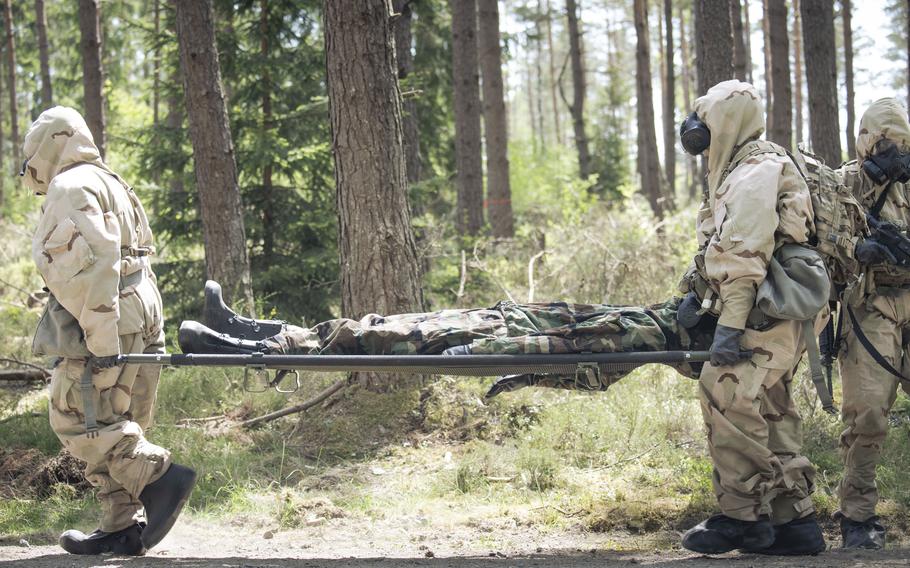 Three soldiers in protective gear and gas masks, two of them carrying a stretcher with a dummy.