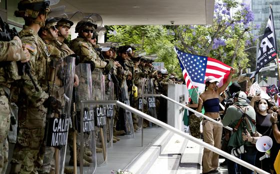A line of Guard members stand outside a federal building while a protestor in front of them holds an American flag.