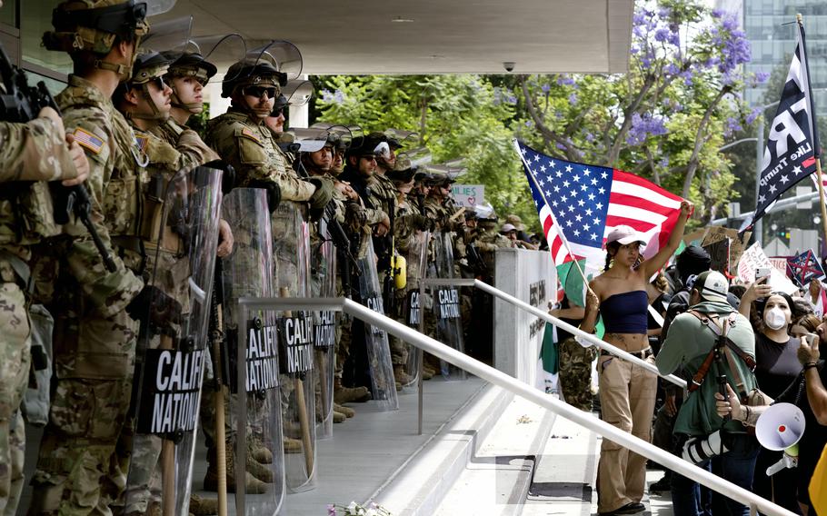 A line of Guard members stand outside a federal building while a protestor in front of them holds an American flag.