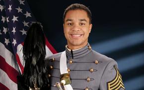 An army cadet stands while holding a ceremonial sword and hat, with a U.S. flag on the left.