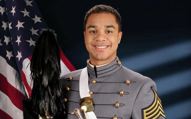 An army cadet stands while holding a ceremonial sword and hat, with a U.S. flag on the left.