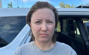 A woman with brown hair and wearing a gray shirt, seen from the neck up, stands next to a car.
