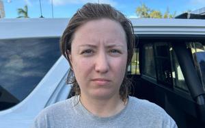 A woman with brown hair and wearing a gray shirt, seen from the neck up, stands next to a car.