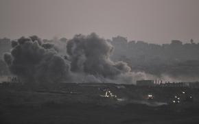 Smoke rises over damaged buildings in the foreground, with a hazy skyline of damaged buildings in the background.