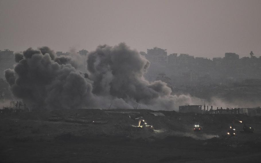 Smoke rises over damaged buildings in the foreground, with a hazy skyline of damaged buildings in the background.