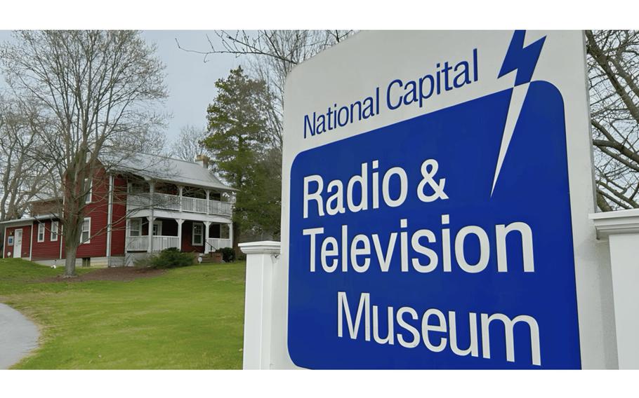 A closeup of a sign that reads “National Capital Radio & Television Museum” with a farmhouse in the background.