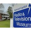 A closeup of a sign that reads “National Capital Radio & Television Museum” with a farmhouse in the background.