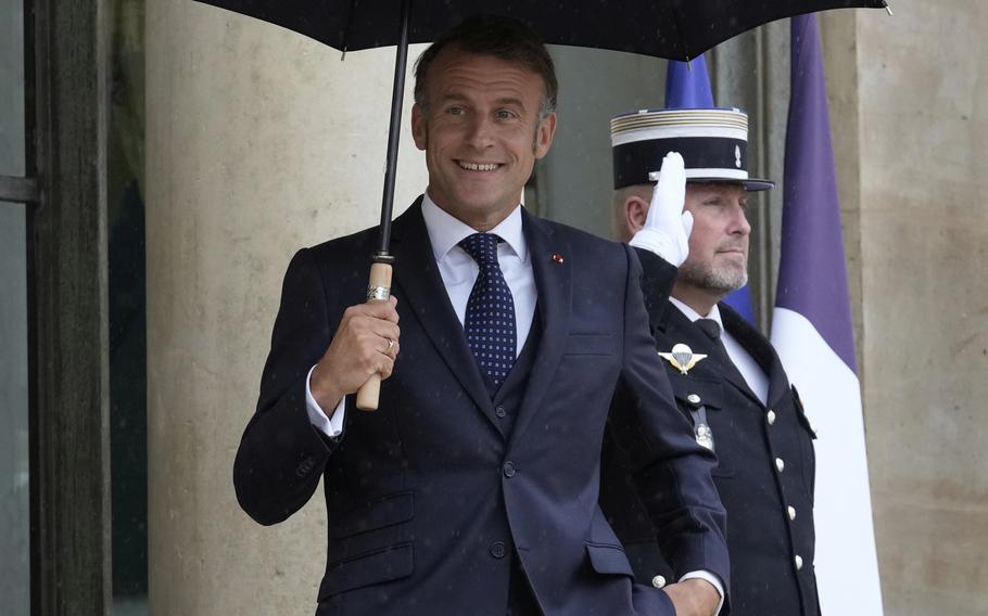 Emmanuel Macron holds an umbrella over his head as he stands outside a government building with a military service member saluting behind him.