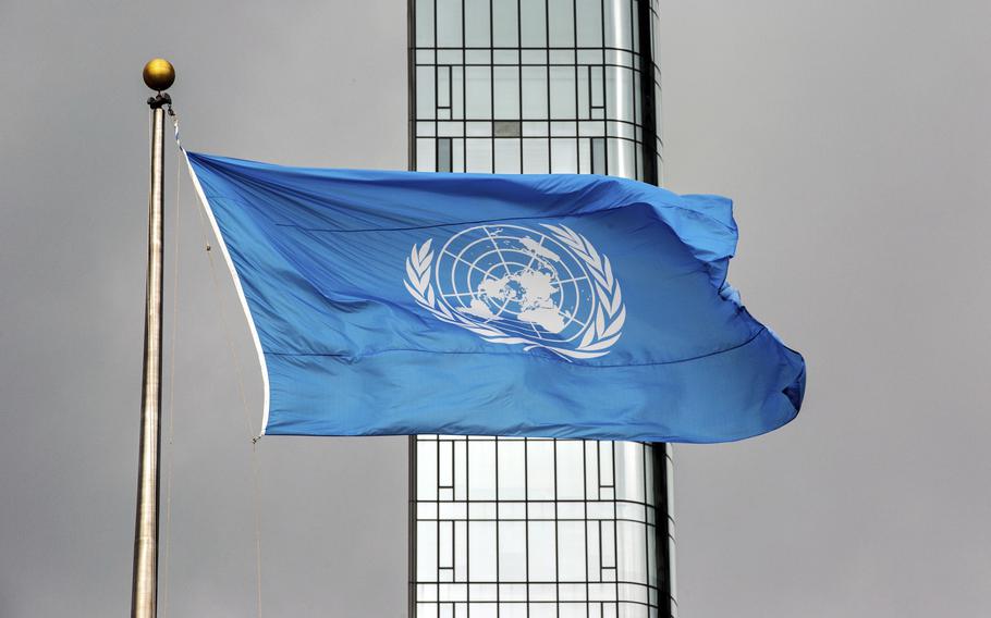 A blue flag with the white emblem of the United Nations flies at the top of a flag pole, with a steel and glass building behind it.