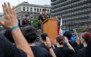 A military officer in dress uniform stands at a podium on an outdoor stage with his right hand raised as recruits in black shirts stand in the foreground with their hands raised to take an enlistment oath.
