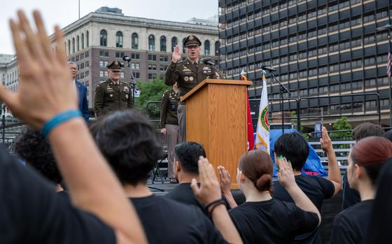 A military officer in dress uniform stands at a podium on an outdoor stage with his right hand raised as recruits in black shirts stand in the foreground with their hands raised to take an enlistment oath.