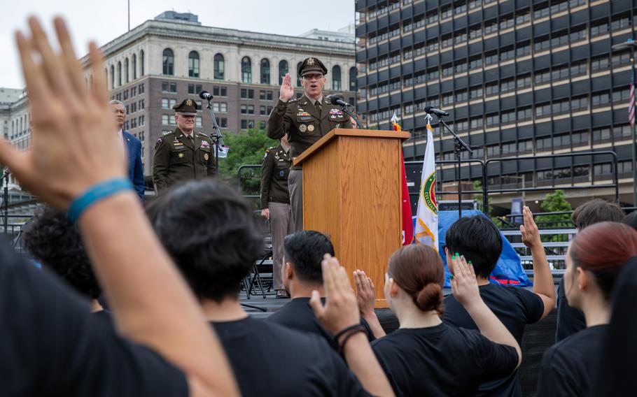 A military officer in dress uniform stands at a podium on an outdoor stage with his right hand raised as recruits in black shirts stand in the foreground with their hands raised to take an enlistment oath.