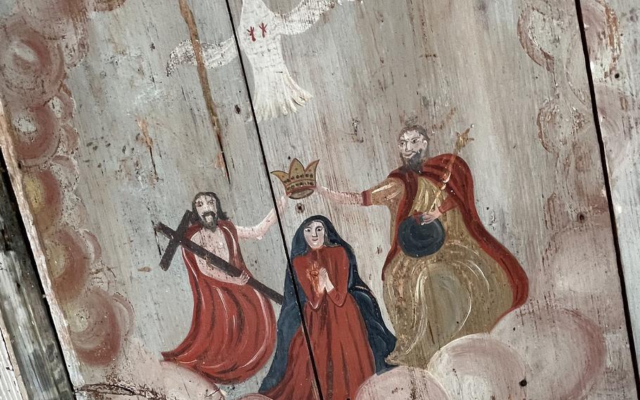 A rustic religious painting on wood depicting the Coronation of the Virgin Mary. She kneels in prayer as Jesus and God the Father hold a crown above her head, with a dove symbolizing the Holy Spirit above them, all surrounded by clouds.