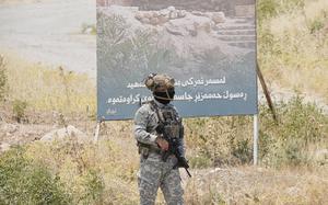 A man in camouflage and holding a rifle walks near a billboard in a grassy area.