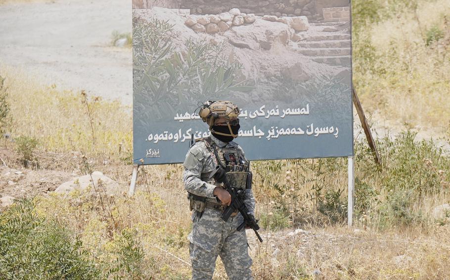 A man in camouflage and holding a rifle walks near a billboard in a grassy area.