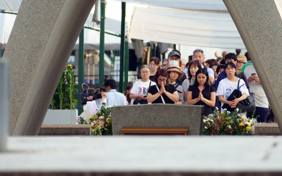 A group of people clasp their hands in prayer in front of a memorial honoring atomic bomb victims.