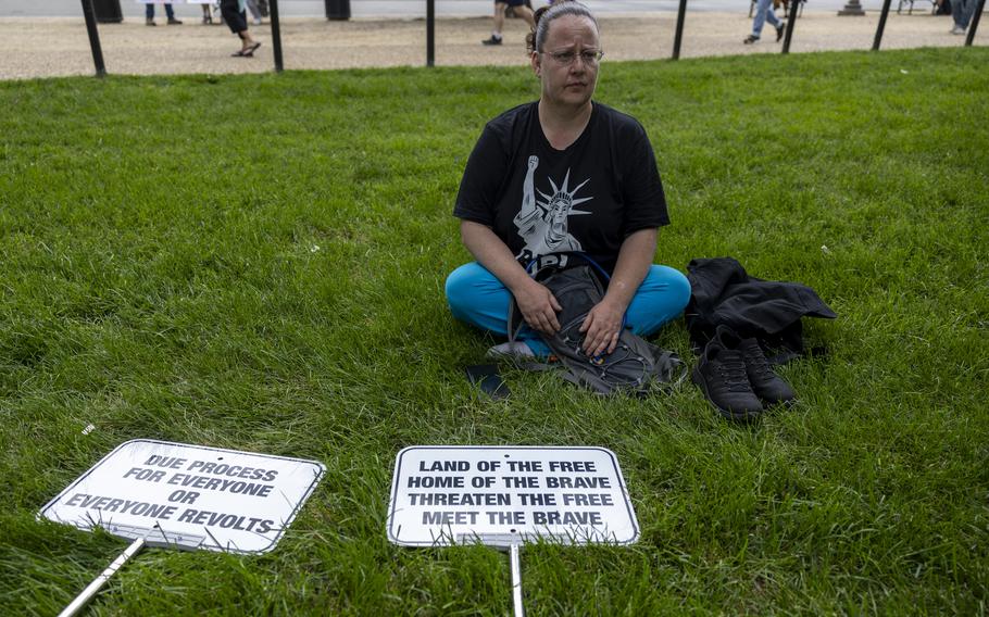 A woman wearing a black shirt depicting the Statue of Liberty with a raised fist sits on a patch of grass with two protest signs lying in front of her.