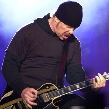 A man in a black beanie and black clothes plays his guitar.