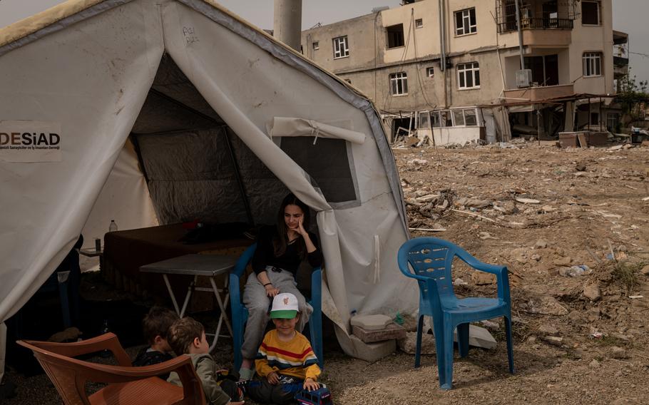 A young woman sits with children in front of their tent and near their former home in Samandag. 