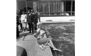 A black and white photo of a barefoot woman sitting by a pool, while other people in formal clothes stand behind her.