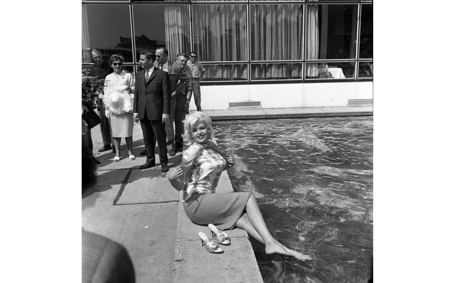 A black and white photo of a barefoot woman sitting by a pool, while other people in formal clothes stand behind her.