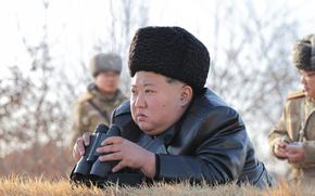 Kim Jong Un, wearing a hat and leather jacket, holds a pair of binoculars as he crouches in a grassy area.