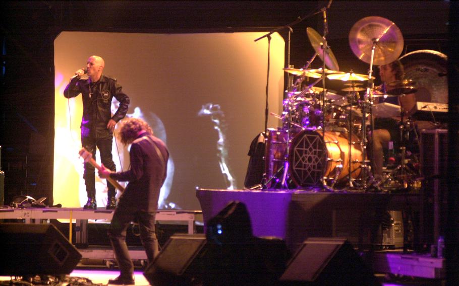 Three men seen on stage, one singing, one playing a guiltar and another on the drums.
