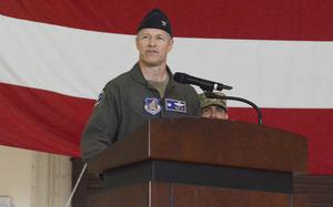 A man in a military flight suit and hat speaks into a microphone while standing at a podium in front of a large U.S. flag.