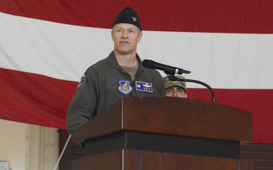 A man in a military flight suit and hat speaks into a microphone while standing at a podium in front of a large U.S. flag.