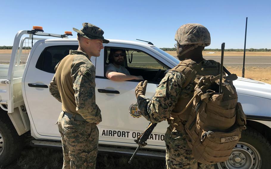 Two military personnel in camouflage uniforms stand beside a white truck marked “Airport Operations” on an airfield. They are conversing with a third person who sits in the vehicle’s driver seat.
