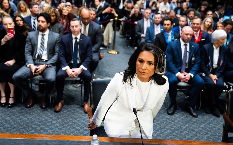 Tulsi Gabbard in a white suit, halfway off her chair as she either prepares to sit down or stands up.