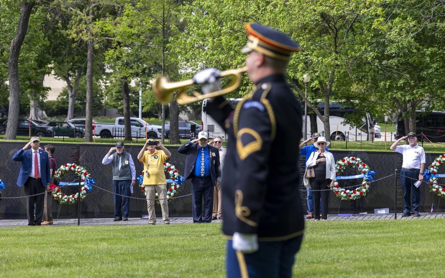 A military bugler in dress uniform performs on a grass field as others watch from along a black memorial wall.